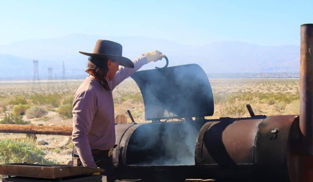 Cowboy lifting the lid of a barbecue smoker, surrounded by a desert landscape, showcasing outdoor BBQ experience related to Covered Wagon Tours in Palm Springs.