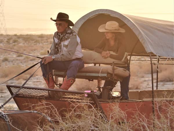 Man and woman seated in a covered wagon, wearing cowboy hats and western attire, enjoying a scenic view in the desert landscape during sunset, representing the authentic experience of Covered Wagon Tours in Palm Springs.