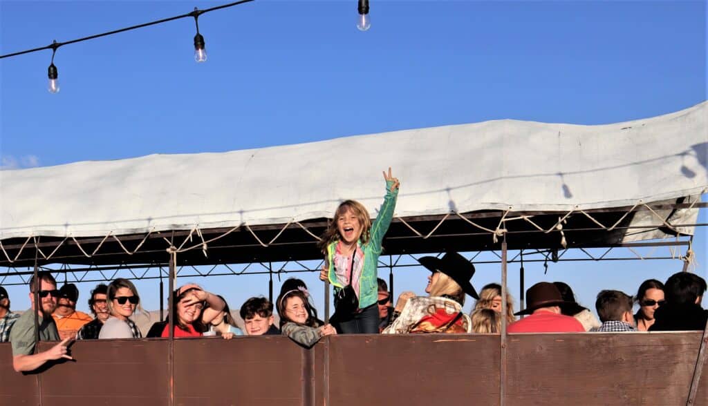 Child joyfully celebrating on a mule-drawn covered wagon surrounded by smiling families, with a clear blue sky and festive lights above, highlighting the fun of Covered Wagon Tours in Palm Springs.